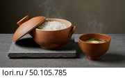 Steaming Rice and Soup Delight: A culinary scene showcasing a traditional Japanese aesthetic with freshly steamed rice in a clay pot, served alongside a complementary bowl of soup. Стоковое AI-изображение, AI-художник Лариса Пономарева / Фотобанк Лори