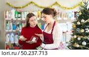 Friendly saleswoman in maroon apron helping teenage girl select makeup palette in Christmas decorated shop, offering advice and guidance. Стоковое видео, видеограф Яков Филимонов / Фотобанк Лори