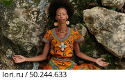 African American woman in a vibrant dress meditates peacefully against a rocky backdrop. Стоковое AI-изображение, AI-художник Данила Большаков / Фотобанк Лори