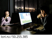 Young adult asian businessman and woman drinking hot cofee while working late at night in their office with desktop computer and laptop. Using as hard working and working late concept. Стоковое фото, агентство Ingram Publishing / Фотобанк Лори