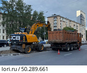 Moscow, Russia - June 23, 2025: Yellow JCB excavator loading asphalt debris into an orange dump truck during roadwork on Budennogo Ave. Редакционное фото, фотограф Софья Соломонова / Фотобанк Лори