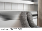 Legs in knitted socks warming over electric heater. Frozen woman wearing a warm woolen socks freezing for winter cold. Discomfort spending time at home. Girl warming her feet on modern radiator. Стоковое фото, фотограф Марина Демидюк / Фотобанк Лори