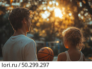 Warm outdoor scene in the rays of the setting sun. A man and a child stand together on a basketball court, holding a ball and looking into the distance, as if preparing for a game. The sunlight softly illuminates their hair, creating an atmosphere of comfort, trust and family closeness. The scene symbolizes moments of parental support, spending time together and the importance of communication between generations. Стоковое AI-изображение, AI-художник Анна Кривицкая / Фотобанк Лори