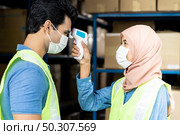 Islam asian warehouse worker taking temperature to worker before getting in factory after reopening from city lockdown from COVID-19 coronavirus pandemic in distribution center environment. Side view. Стоковое фото, агентство Ingram Publishing / Фотобанк Лори