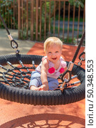 A joyful baby plays on a swing, surrounded by vibrant colors and greenery on a sunny day. Стоковое фото, фотограф Данила Большаков / Фотобанк Лори