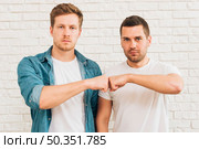 portrait two male friends bumping fist standing against white brick wall. Стоковое фото, агентство Ingram Publishing / Фотобанк Лори