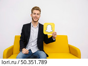 portrait young man sitting yellow sofa holding snapchat icon. Стоковое фото, агентство Ingram Publishing / Фотобанк Лори