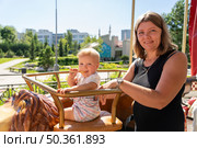 A mother smiles as her baby plays with a snack while sitting on a carousel horse in the park on a bright, sunny day. Стоковое фото, фотограф Данила Большаков / Фотобанк Лори