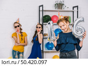 birthday girl showing numeral 16 foil silver balloons with her two friends blowing party blower. Стоковое фото, агентство Ingram Publishing / Фотобанк Лори