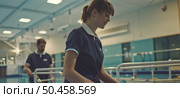 A focused young female nurse in blue scrubs proceeds through a hospital corridor, with a male colleague in the background. Стоковое AI-изображение, AI-художник Данила Большаков / Фотобанк Лори