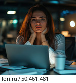 A young woman is sitting in front of a laptop in an office late at night. Her face expresses fatigue, stress and emotional tension. She holds her head in her hands, there are documents scattered on the table and a paper cup with a drink. The atmosphere of the scene conveys a feeling of overload and fatigue from work, especially in the conditions of overtime and deadlines. This photo is suitable for illustrating themes related to burnout, office life, stress at work and emotional health. Стоковое AI-изображение, AI-художник Анна Кривицкая / Фотобанк Лори