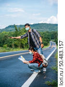 Female tourists sit and look at the map, male tourists pretending to hitchhike. Both wear masks and are on the side of the road. Стоковое фото, агентство Ingram Publishing / Фотобанк Лори