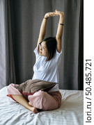 A woman in a white shirt sitting on the bed and raising both arms. Стоковое фото, агентство Ingram Publishing / Фотобанк Лори