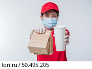 Image of a happy young delivery man in red cap blank t-shirt uniform face mask gloves standing with empty brown craft paper packet isolated on light gray background studio, Service during pandemic coronavirus virus covid-19 concept. Стоковое фото, агентство Ingram Publishing / Фотобанк Лори