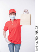 Image of a happy young delivery man in red cap blank t-shirt uniform face mask gloves standing with empty brown craft paper packet isolated on light gray background studio, Service during pandemic coronavirus virus covid-19 concept. Стоковое фото, агентство Ingram Publishing / Фотобанк Лори