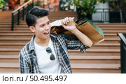 Portrait Handsome young man grinning as he holds a paper bag and shows his credit card on the stairs of a department store. New Normal lifestyle and shopping. Стоковое фото, агентство Ingram Publishing / Фотобанк Лори