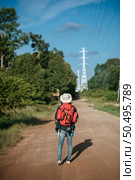 Rear view of young man traveler with backpack wearing sombrero and holding paper map in hand, He walking  and look forward, has high voltage pole and the overgrown bushes beside on the street. Стоковое фото, агентство Ingram Publishing / Фотобанк Лори