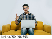 Young handsome man wearing eyeglasses with new normal, sitting on sofa with thump up, Heworks from home on laptop computer while self-isolation and quarantine covid-19 coronavirus outbreak. Стоковое фото, агентство Ingram Publishing / Фотобанк Лори