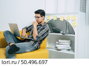 Young handsome man wearing eyeglasses with new normal, sitting on sofa and works from home on laptop computer while self-isolation and quarantine covid-19 coronavirus outbreak. Стоковое фото, агентство Ingram Publishing / Фотобанк Лори