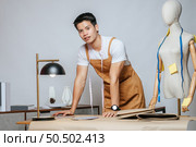 Portrait Young male fashion designer working with fabric and clothing sketches at the studio at home, full of tailoring tools, model and equipment on table. Стоковое фото, агентство Ingram Publishing / Фотобанк Лори