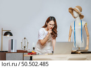 Seriously young Asian female fashion designer or costume use laptop computer while design a beautiful sombrero and taking on mannequin. Стоковое фото, агентство Ingram Publishing / Фотобанк Лори
