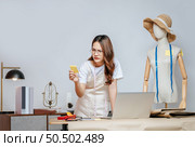 Asian young pretty female fashion designer stylish wearing eyeglasses standing and serious with paper not during working at fashion studio, full of tailoring tools and equipment. Стоковое фото, агентство Ingram Publishing / Фотобанк Лори
