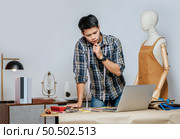 Seriously young Asian fashion designer or tailor working with laptop computer to search idea or online orders, full of tailoring tools with mannequin and equipment on the desk in the studio. Стоковое фото, агентство Ingram Publishing / Фотобанк Лори