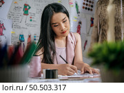 Selective focus, beautiful young designer female or tailor sketching fashion clothes on paper, full of sewing tools on table, sketch paper on board in blured background. Стоковое фото, агентство Ingram Publishing / Фотобанк Лори