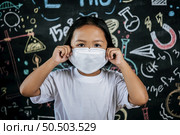 Portrait of a little girl wearing eyeglasses and facemask at school to avoid the spread of coronavirus, new normal with schoolchild,  blure blackboard in background. Стоковое фото, агентство Ingram Publishing / Фотобанк Лори