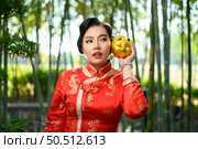 Portrait pretty Asian woman in a Chinese cheongsam posing with piggy bank on her cheek in bamboo forest background, copy space, Chinese New Year concept. Стоковое фото, агентство Ingram Publishing / Фотобанк Лори