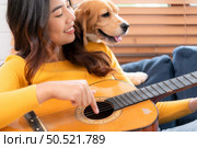On a weekend, A young Asian woman and her beagle puppy are playing the guitar and singing cheerfully together in the living room of a house. Стоковое фото, агентство Ingram Publishing / Фотобанк Лори