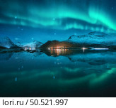 Northern lights over the snowy mountains, sea, fjord, reflection in water at winter starry night. Lofoten, Norway. Aurora borealis and snow covered rocks. Landscape with polar lights, city lights, sky. Стоковое фото, агентство Ingram Publishing / Фотобанк Лори