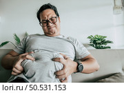 Fat man in glasses sitting on sofa and show obesity with stress, Funny overweight man usd hands squeeze his belly fat, copy space. Стоковое фото, агентство Ingram Publishing / Фотобанк Лори