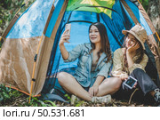 front view Young Asian pretty woman and her girlfriend sitting at front of tent, use mobile phone take photo during camping in forest with happiness together. Стоковое фото, агентство Ingram Publishing / Фотобанк Лори