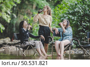 Group of young asian women drink beer in their chairs and soaked their feet in the stream while camping in the nature park, They are enjoy to talking and laugh fun together. Стоковое фото, агентство Ingram Publishing / Фотобанк Лори