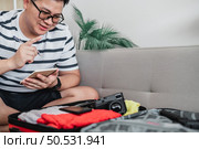 Asian man glasses is preparing clothes in suitcases. He’s choosing clothes, travel documents, the itinerary for a solo trip, and check the checklist. Travel, holiday and vacation concept. Стоковое фото, агентство Ingram Publishing / Фотобанк Лори