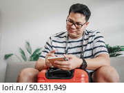 Asian man glasses is preparing clothes in suitcases. He’s choosing clothes, travel documents, the itinerary for a solo trip, and check the checklist. Travel, holiday and vacation concept. Стоковое фото, агентство Ingram Publishing / Фотобанк Лори