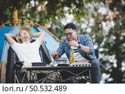 Asian relaxed man using smartphone and waiting for his friend cooking with tent background in camping. Cooking set front ground. Outdoor cooking, traveling, camping, lifestyle concept. Стоковое фото, агентство Ingram Publishing / Фотобанк Лори
