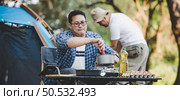 Asian man preparing pitch a tent in camping while the other man was preparing food. in camping. Cooking set front ground. Outdoor cooking, traveling, camping, lifestyle concept. Стоковое фото, агентство Ingram Publishing / Фотобанк Лори