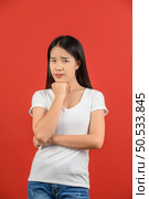 Portrait of distrustful young asian woman in white t-shirt hand chin isolated on red background. Expression and lifestyle concept. Стоковое фото, агентство Ingram Publishing / Фотобанк Лори