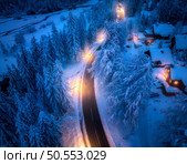 Aerial view of road in fairy town in snow, forest, houses with street lights at night in winter. Top view of village, rural road, illumination, snowy pine trees at dusk in Kranjska Gora, Slovenia . Стоковое фото, агентство Ingram Publishing / Фотобанк Лори
