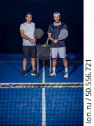 Full length portrait of two sporty young men in sportswear standing on indoor court with paddle rackets in hands after match. Стоковое фото, агентство Ingram Publishing / Фотобанк Лори