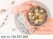 Quail eggs in a ceramic bowl with a peach fuzz pink colored scarf and bird feathers. Wooden table painted white. Background with elements suggestive of the Easter season. Стоковое фото, агентство Ingram Publishing / Фотобанк Лори