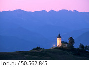 A church on a hill at sunset with mountain ranges in the background. Peaceful and serene atmosphere. Illuminated church against high alpine mountains and pink sky at twilight in Slovenia. Landscape. Church on a hill at dusk with mountain ranges in the background. Стоковое фото, агентство Ingram Publishing / Фотобанк Лори