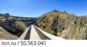 A wooden walkway leading to a dam, surrounded by rugged terrain under a clear blue sky. Guarda, Portugal. Стоковое фото, агентство Ingram Publishing / Фотобанк Лори