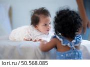 A small girl stood at the foot of the bed playing with the younger sibling carried by his mother and smiling with affection, In a bedroom surrounded by warmth and family. Стоковое фото, агентство Ingram Publishing / Фотобанк Лори