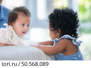 A small girl stood at the foot of the bed playing with the younger sibling carried by his mother and smiling with affection, In a bedroom surrounded by warmth and family. Стоковое фото, агентство Ingram Publishing / Фотобанк Лори
