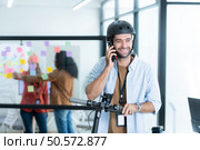 Portrait of smiling businessman talking on mobile phone while colleague using scooter in office. Стоковое фото, агентство Ingram Publishing / Фотобанк Лори