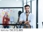 Portrait of smiling businessman riding electric scooter in office with colleague in background. Стоковое фото, агентство Ingram Publishing / Фотобанк Лори