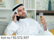 Portrait of a smiling Arabian man holding a glass of champagne and using mobile phone at home. Стоковое фото, агентство Ingram Publishing / Фотобанк Лори